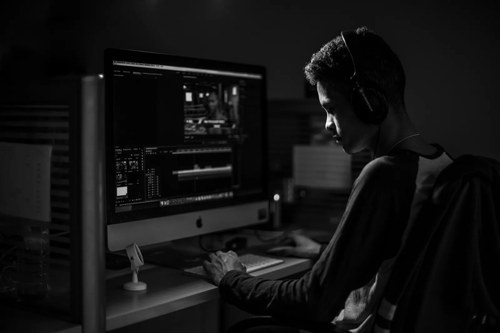 A man wearing headphones working on video editing on a computer in a dimly lit office.