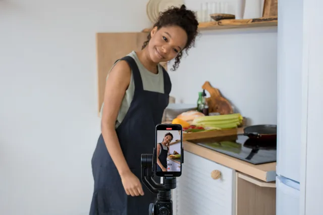 Smiling young woman films a cooking video at home using a smartphone in her kitchen. UGC hooks and opening scenes